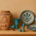 A gorgeous collection of Egyptian ceramics on a wooden bench against a beige background. Features include ancient Egyptian pottery with hand-drawn motifs, handpainted Fayoum pottery plates and jugs, an small Egyptian Faience pieces, evoking a rustic, cultural ambiance.