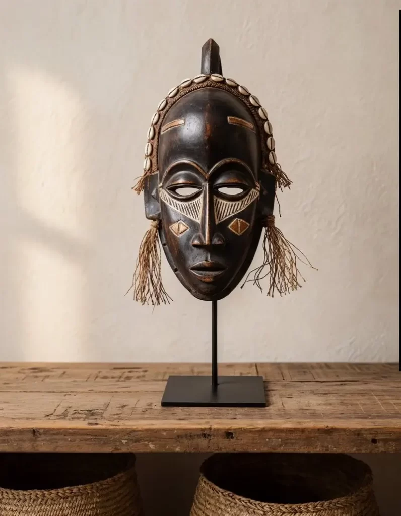 A dark-toned, hand-carved African artifacts mask featuring intricate white geometric patterns on the cheeks and forehead, adorned with a cowrie shell headband and natural fiber tassels, displayed on a black metal stand.