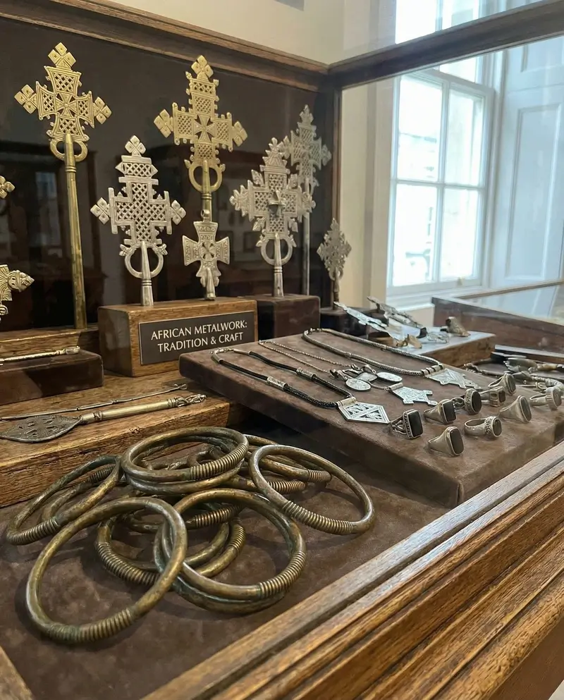 A museum-style display case featuring various metal African artifacts, including ornate Ethiopian processional crosses, coiled bronze armbands, and silver Tuareg rings.