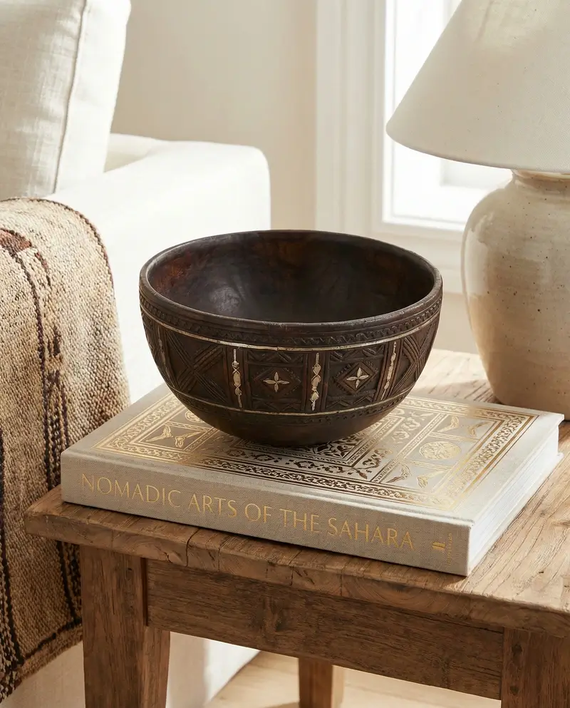 A hand-carved dark wood Tuareg bowl with intricate geometric silver inlays, displayed on a coffee table book titled "Nomadic Arts of the Sahara."
