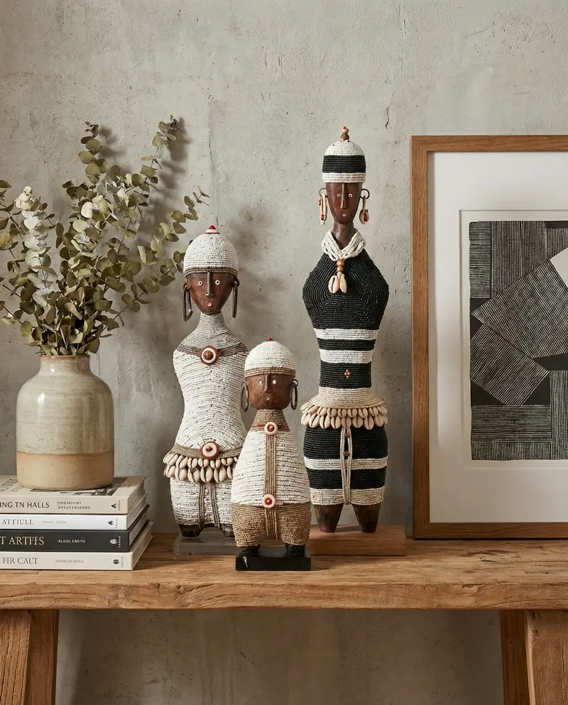 A trio of traditional Namji dolls of varying heights, intricately adorned with white and black beadwork and cowrie shell belts, styled on a rustic wooden console. These African artifacts are celebrated for their unique silhouettes and craftsmanship.