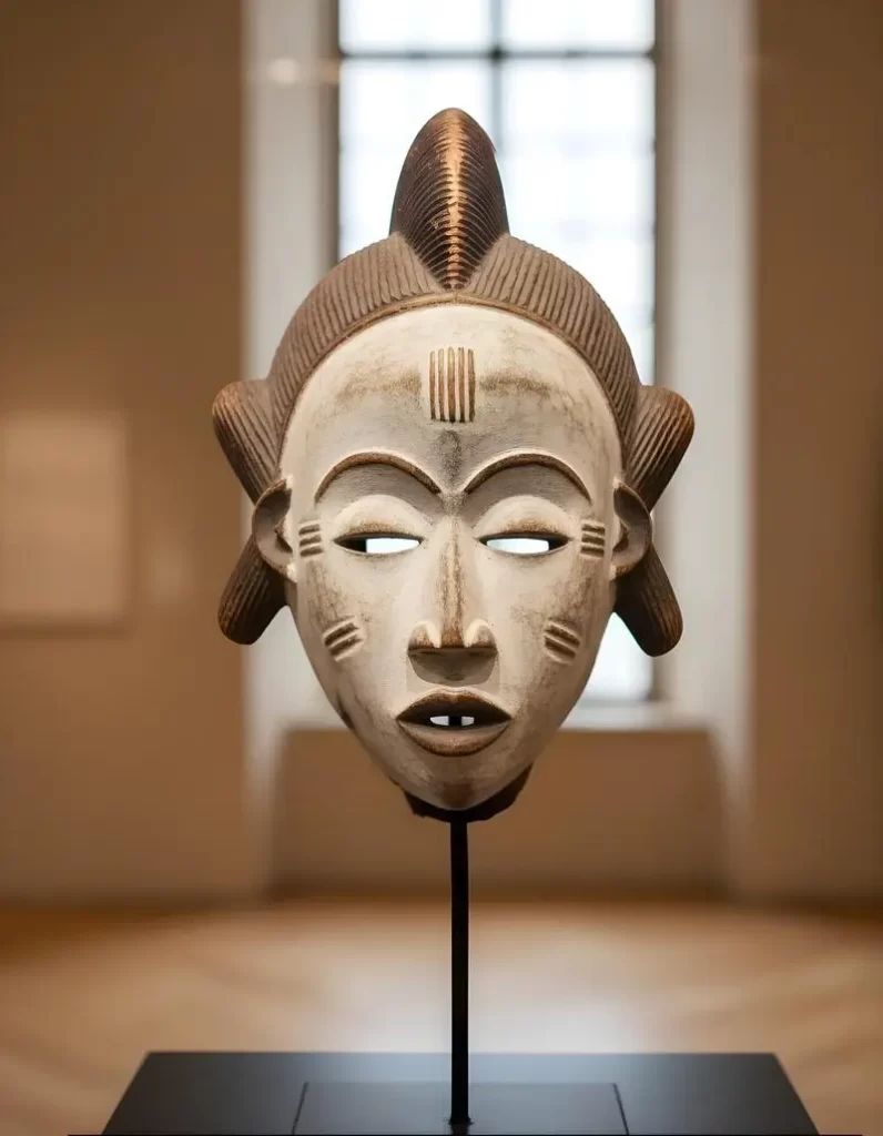 A traditional Punu mask from Gabon, characterized by a white-pigmented face, high-arched eyebrows, and a distinctive tri-lobed hairstyle, presented as a museum-quality sculpture on a minimalist stand.