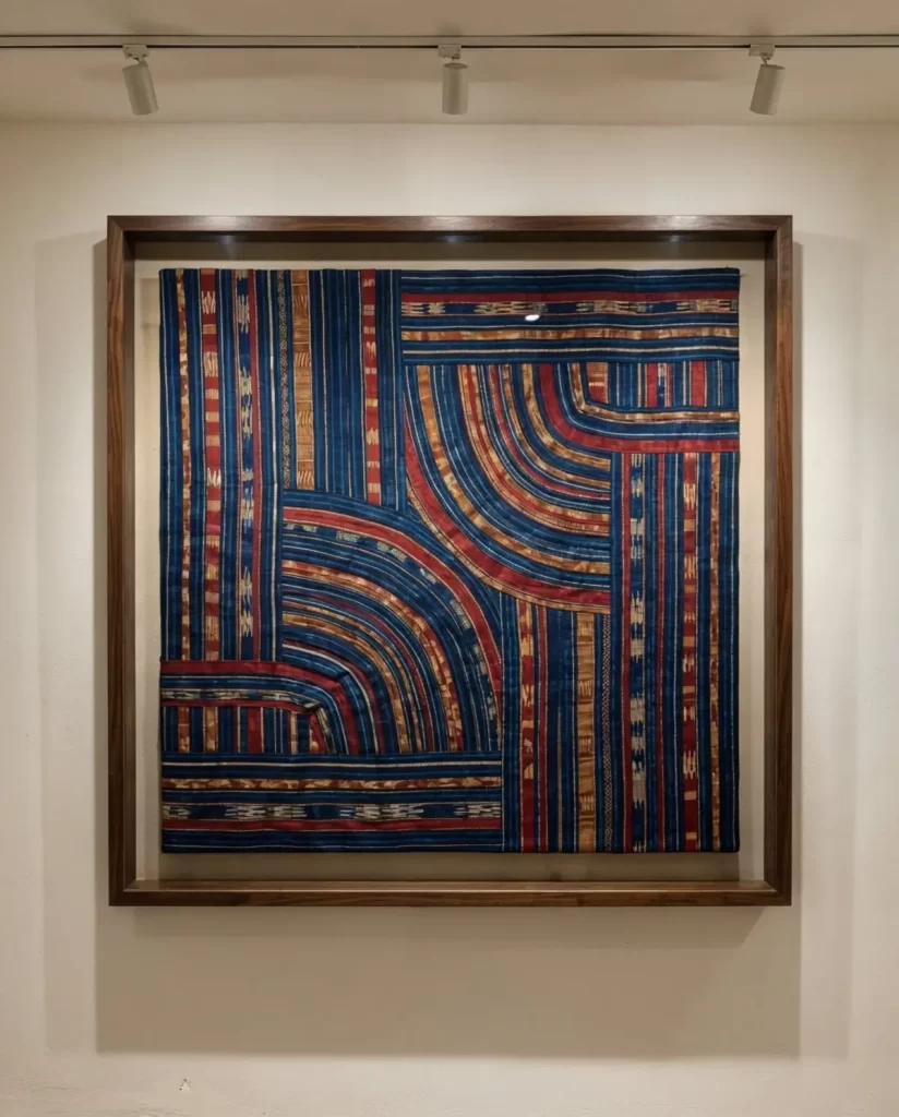 Framed contemporary African textile art with a pattern of curved and straight lines in deep blue, rust red, and golden-tan, hanging on a white wall.