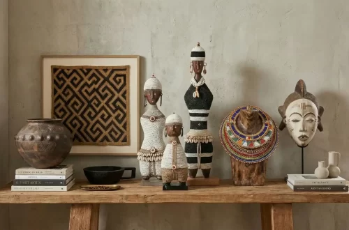 A gorgeous display of African artifacts: left to right: a Zulu pot placed on a stack of books, framed kuba cloth on the wall, an Ethiopian Gurage bowl, 3 namji dolls at the center, a Maasai bead collar on a wood bust and a Punu mask on a stand