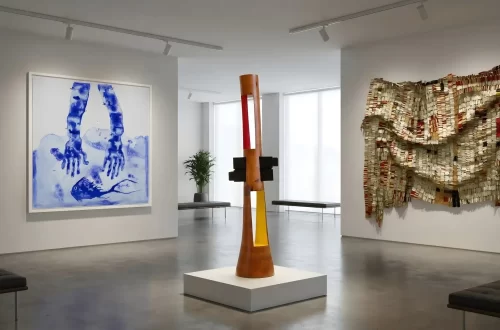 A bright, modern art gallery space showcasing works by African artists, including a large blue and white painting of hands, a tall wooden and glass sculpture, and a textural wall hanging made of layered materials.
