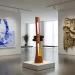 A bright, modern art gallery space showcasing works by African artists, including a large blue and white painting of hands, a tall wooden and glass sculpture, and a textural wall hanging made of layered materials.