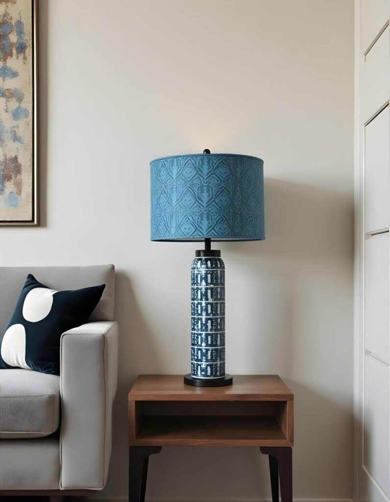 A tall, slender table lamp with a blue and white patterned ceramic base and a matching blue fabric drum shade featuring an intricate African-inspired motif. The lamp sits on a mid-century modern wooden side table next to a light gray sofa. A black throw pillow with large white circles rests on the sofa, and a portion of an abstract painting is visible on the wall to the left.