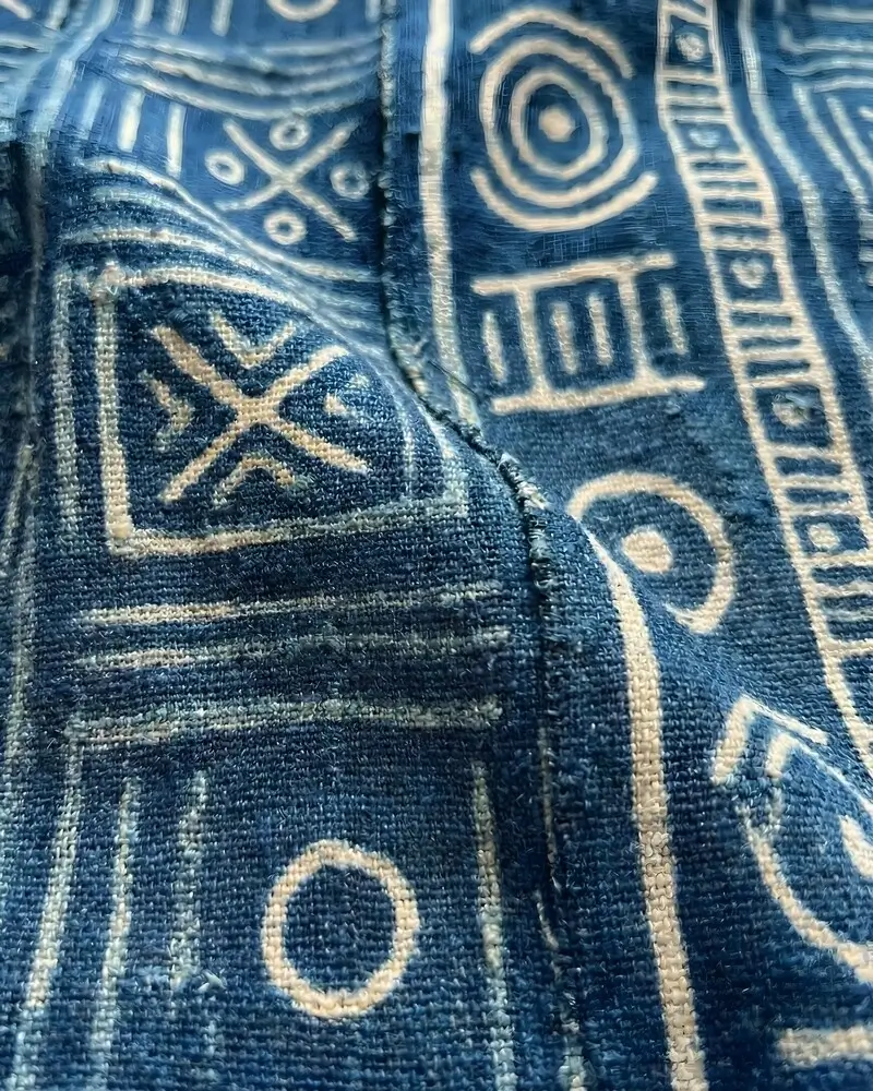 A close-up of a heavy-weave African indigo fabric featuring cream-colored symbolic motifs, including circles, crosses, and linear borders, showcasing the artisanal hand-dyed quality.