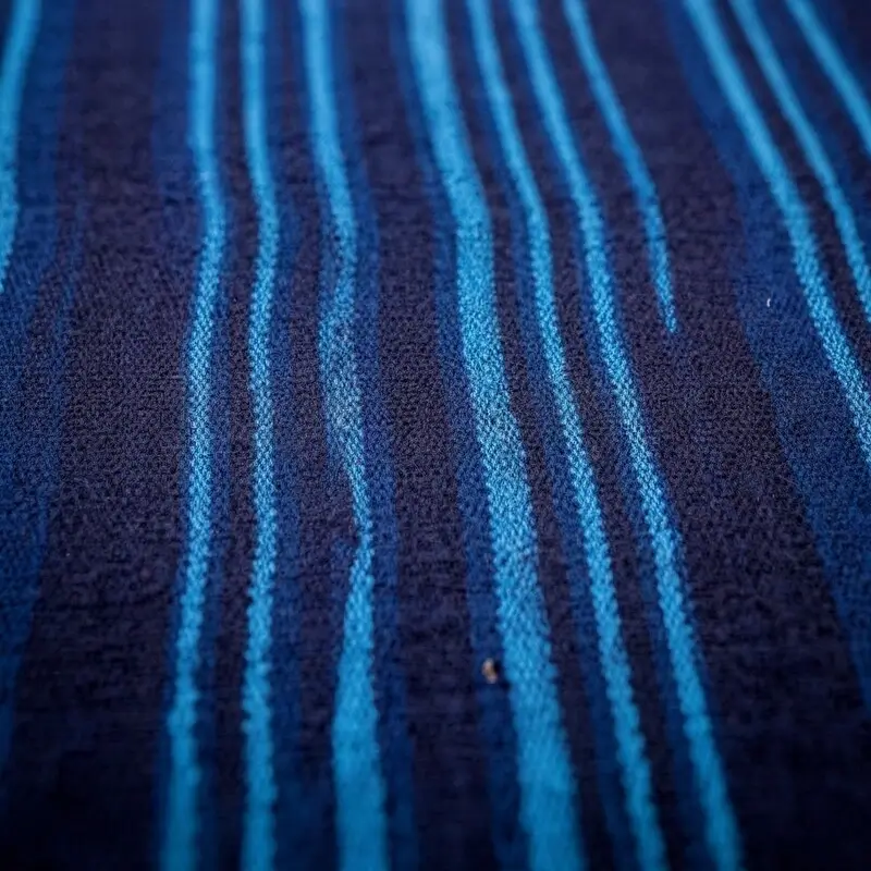 A macro shot of Leppi fabric from Guinea, displaying a series of vertical, vibrant cyan stripes woven into a dark navy blue base, highlighting the fine texture of the weave.