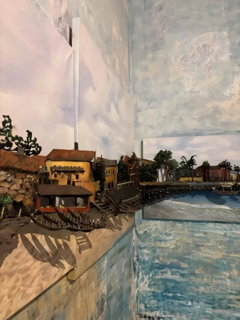 A 3D diorama or wall sculpture by Loman Pawlitschek depicting a coastal village. Tiny, detailed houses and traditional pirogue boats are mounted over a painted background of a cloudy sky and turquoise ocean water.
