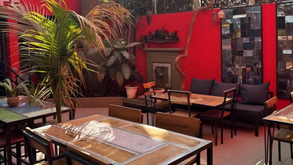 A close-up of an outdoor seating area with a bold red wall. The space features wooden tables with artistic inlays, black cushioned benches, and lush tropical plants under warm, natural light.