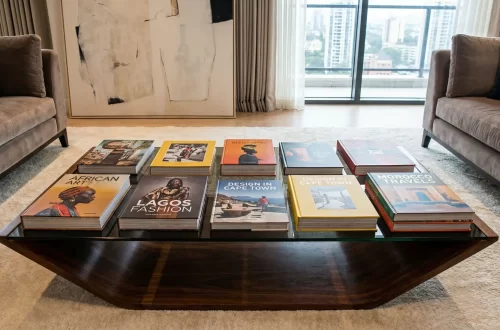 A wide-angle, eye-level shot of a modern living room featuring a glass-topped wooden coffee table arranged with several decorative African books. The books, which showcase vibrant covers with titles like "African Art," "Lagos Fashion," and "Morocco Travels," are laid out in two neat rows. The room is styled with plush grey velvet sofas, a large abstract painting, and floor-to-ceiling windows offering a city view.