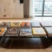 A wide-angle, eye-level shot of a modern living room featuring a glass-topped wooden coffee table arranged with several decorative African books. The books, which showcase vibrant covers with titles like "African Art," "Lagos Fashion," and "Morocco Travels," are laid out in two neat rows. The room is styled with plush grey velvet sofas, a large abstract painting, and floor-to-ceiling windows offering a city view.