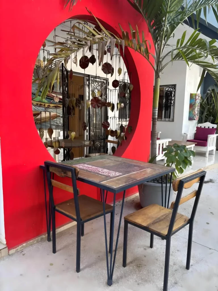 A cozy cafe seating area featuring a wooden table and two chairs in front of a bold red wall with a large circular window. Ornate metal hangings and a potted palm tree add to the artistic, relaxed vibe.