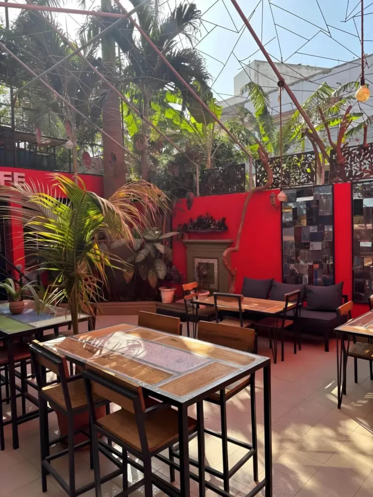 A broad view of the open-air cafe at Loman Art House. A geometric metal framework overhead supports hanging lights, while red walls, green palm trees, and modern wooden furniture create a colorful, creative atmosphere.