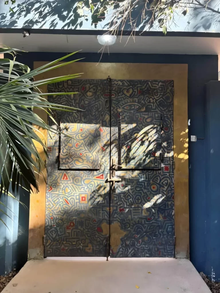 A pair of large, weathered metal doors completely covered in intricate, hand-painted patterns and symbols. The doors are set within a yellow frame against a blue building, with shadows of nearby leaves dancing across the surface.