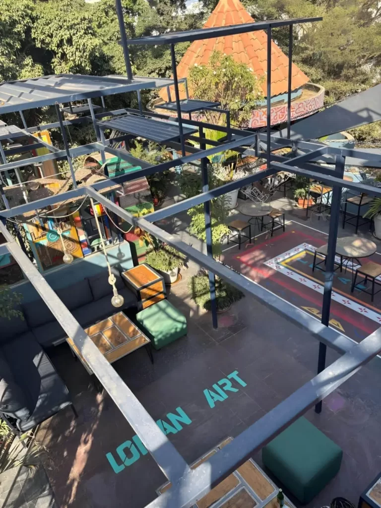 An overhead view of a modern rooftop terrace featuring a dark metal trellis, comfortable black lounge seating, and geometric-patterned rugs on the floor. The words "LOMAN ART" are painted on the ground.