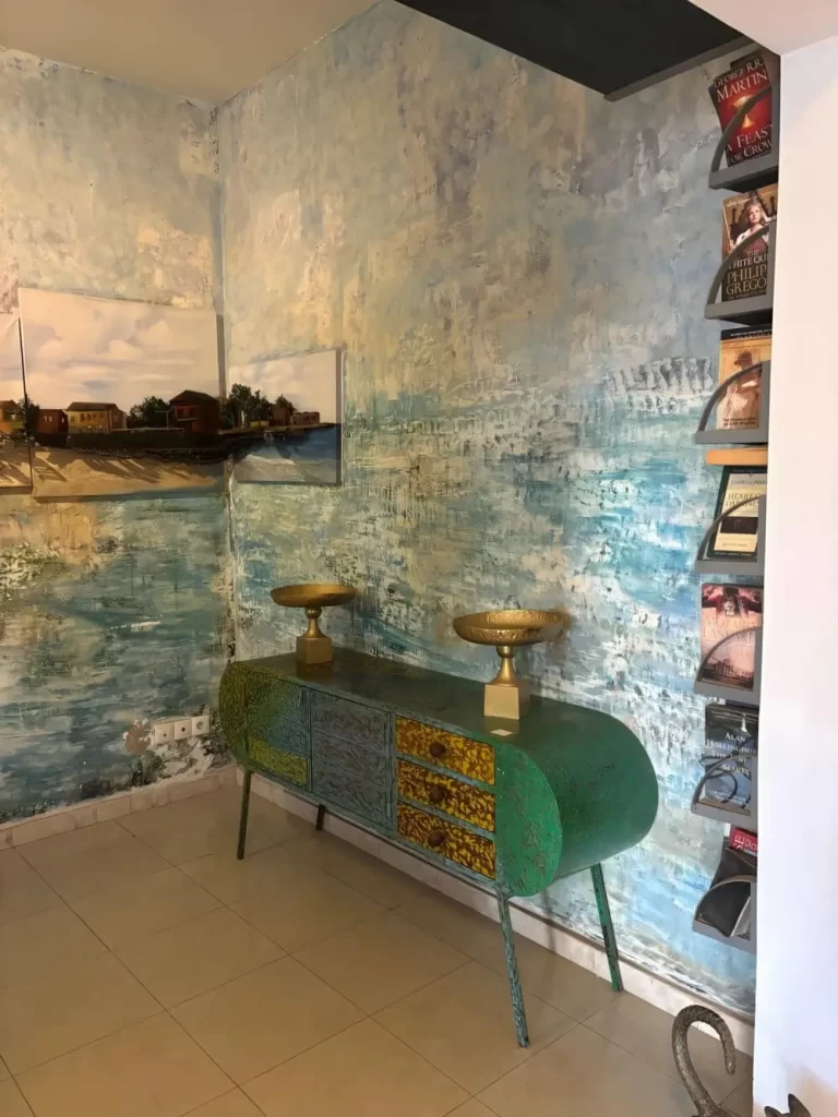 An interior corner showing a wall with a textured blue-and-white mural. A sleek green-and-yellow patterned sideboard sits against the wall, topped with two gold bowls, next to a vertical bookshelf filled with novels.
