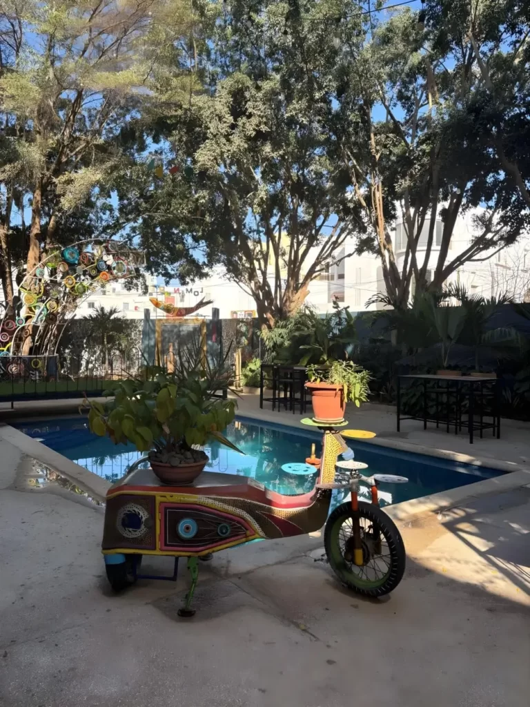 A courtyard scene featuring a swimming pool with a unique sculpture of a motor scooter in the foreground. The scooter is decorated with intricate patterns and used as a planter for lush green vegetation.