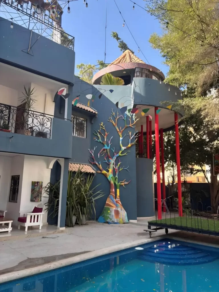 A wide exterior view showing the swimming pool adjacent to the main building. The deep blue facade is decorated with a large painted tree sculpture and hanging colorful pennants, with a small grass-covered bridge crossing the pool area.
