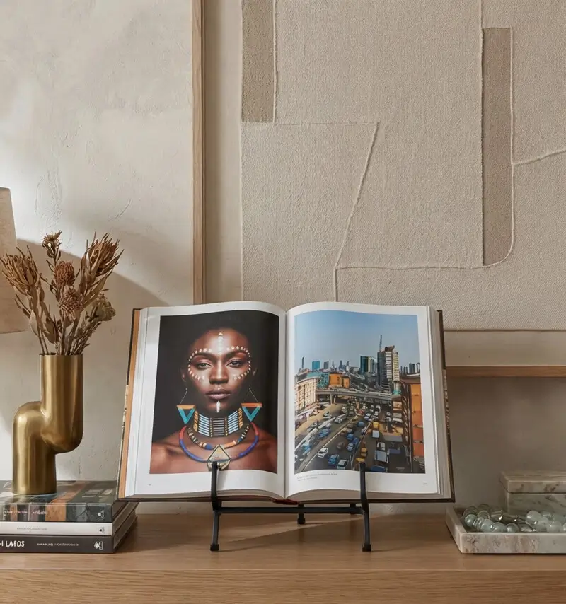 An open coffee table book displayed on a black metal stand atop a wooden console table. Part of a collection of decorative African books, the pages show a striking portrait of a woman in traditional jewelry alongside a bustling African cityscape.