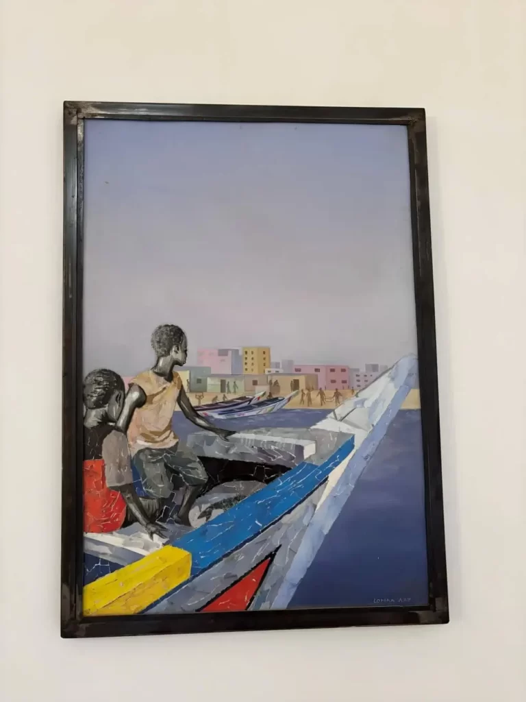A framed painting by Loman Pawlitschek depicting two people in a traditional Senegalese pirogue boat on a deep blue sea, with a coastal town of colorful buildings visible in the distance.