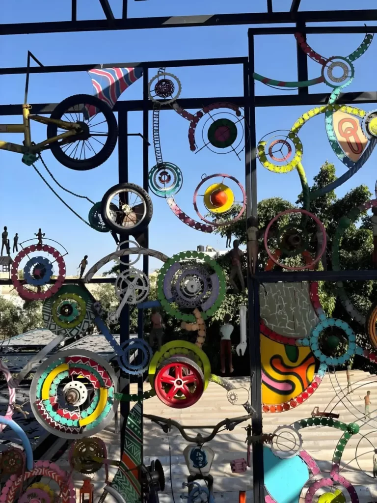 A complex, large-scale outdoor installation made from recycled bicycle parts. Metal frames, gears, and wheels are painted in bright colors and arranged geometrically against a clear blue sky.