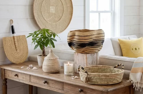 African coastal summer edit: A wooden table displays woven baskets, a potted plant, a lidded jar, a white candle, and seashells. Behind, a neutral sofa with yellow and white pillows and a woven wall hanging complete the cozy, sunlit room.