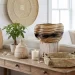 African coastal summer edit: A wooden table displays woven baskets, a potted plant, a lidded jar, a white candle, and seashells. Behind, a neutral sofa with yellow and white pillows and a woven wall hanging complete the cozy, sunlit room.
