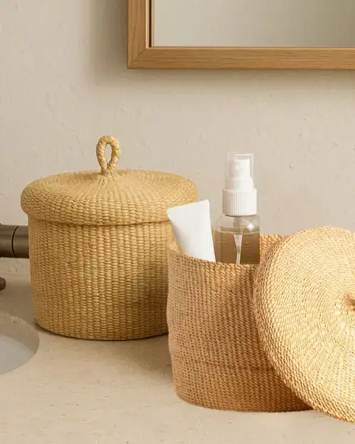 Two woven baskets with lids sit on a bathroom counter. One basket is open, holding a spray bottle and a tube. A mirror and part of a sink are visible in the background.