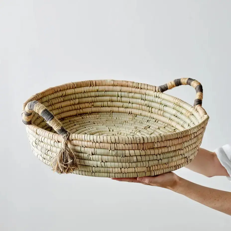 A hand holds a round, woven basket with two handles wrapped in dark and light material. The basket is natural-colored and has a rustic, handmade appearance against a plain background.