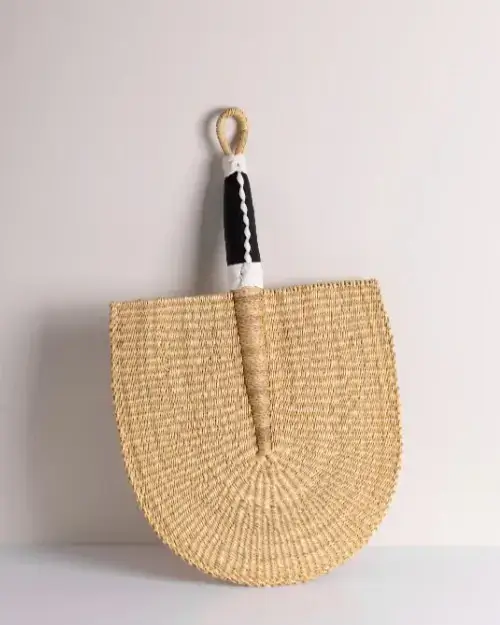A handwoven straw fan with a semicircular shape and a black-and-white wrapped handle, hanging on a light-colored wall.