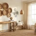 A sunlit coastal grandmother room with a rustic wooden console table, bench, and wicker chair. Woven baskets and ceramic art decorate the wall. Sheer curtains frame a tall window; a jute rug and greenery complete the cozy, natural decor.