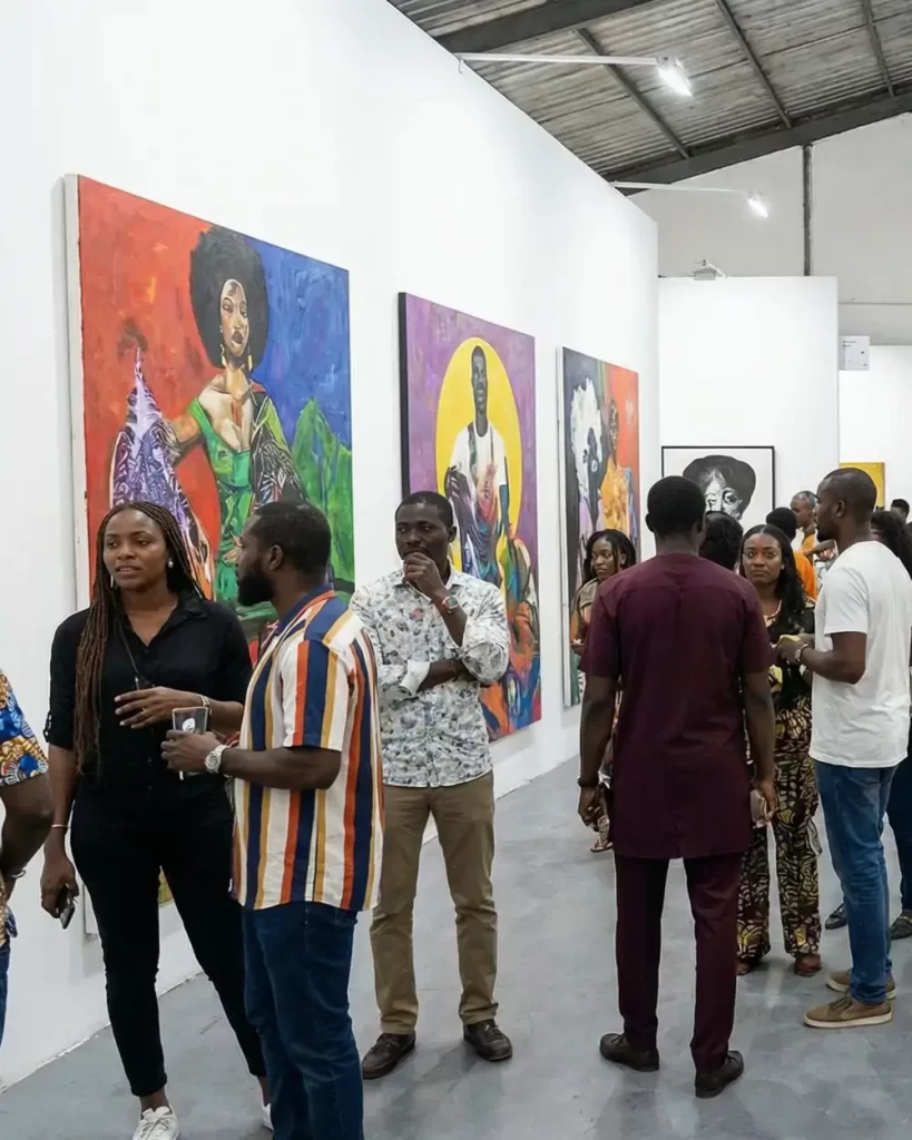 People view colorful portrait paintings at a contemporary African art fair. Several groups are engaged in conversation, and the atmosphere is lively and social. The gallery has a high, industrial ceiling.