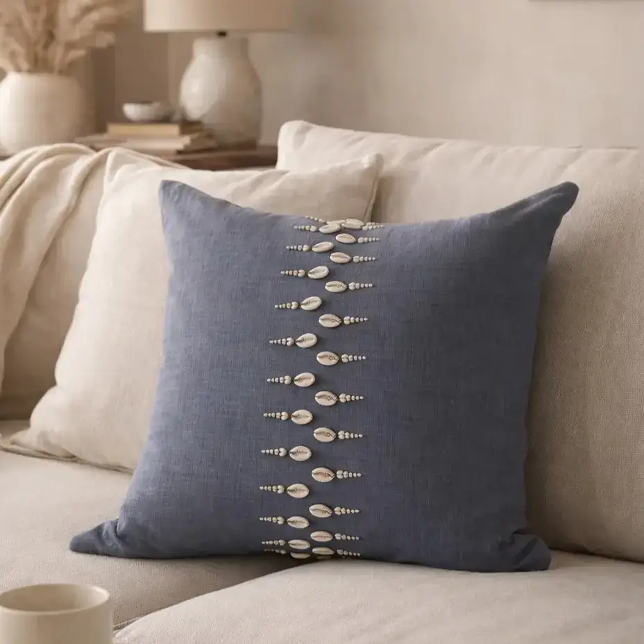 A square blue-gray cushion with a vertical pattern of decorative shells and beads sits on a light beige sofa in a cozy, neutral-toned living room.