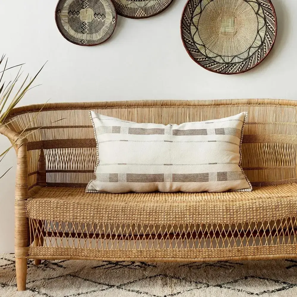 A rattan wicker sofa with a beige and brown patterned cushion sits on a geometric rug. Three decorative woven baskets hang on a white wall above the sofa. A plant is partially visible on the left.