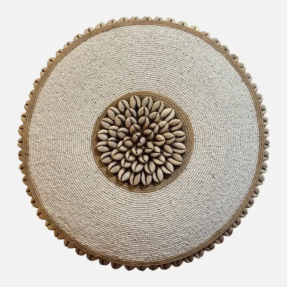 A round decorative object adorned with a central cluster of cowrie shells, surrounded by concentric circles of small white beads and bordered with more cowrie shells, creating a textured, symmetrical design.