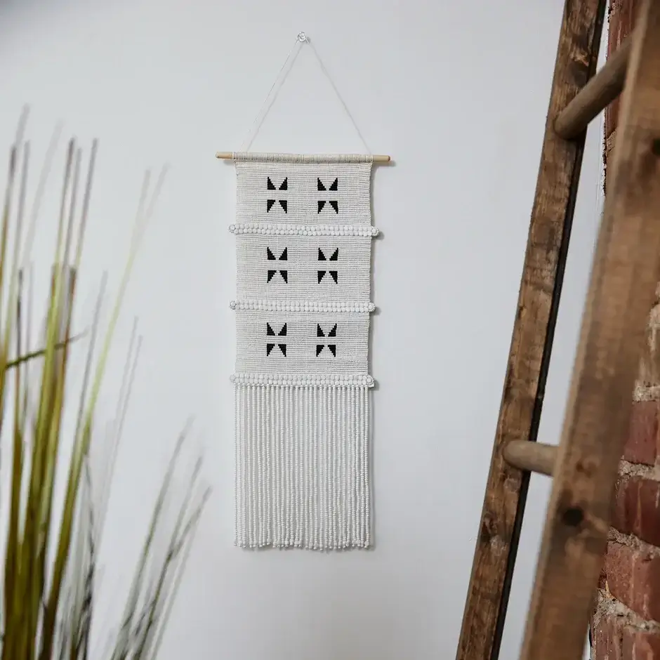 A woven wall hanging with black geometric patterns and long white fringe, displayed on a wooden rod against a white wall near a ladder and tall green plant.