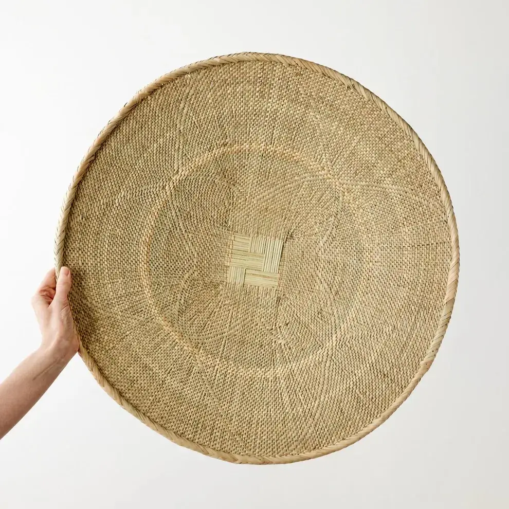 A hand holds up a large, round, woven basket tray made of natural fibers, featuring a textured pattern and a central square design against a plain light background.