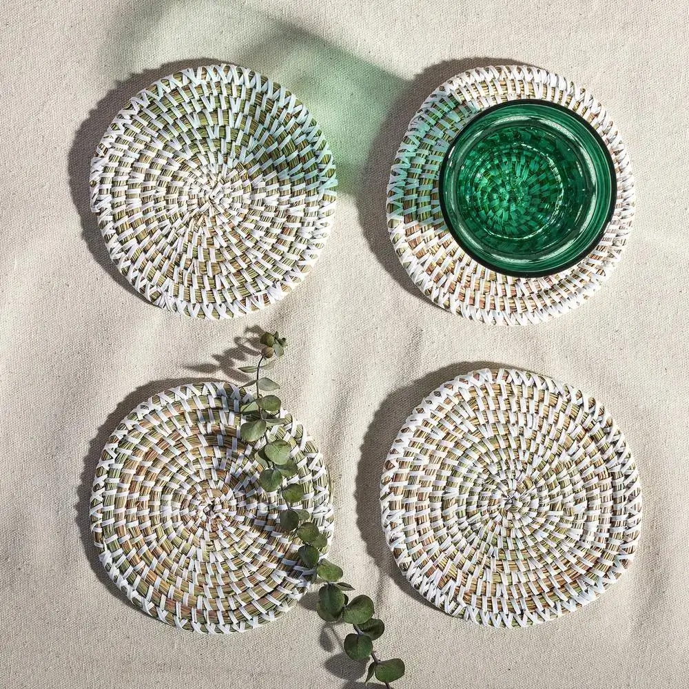 Four round woven coasters are arranged in a grid on a light fabric surface. A green glass cup sits on the top right coaster, while a small eucalyptus branch lies diagonally across the bottom left coaster.
