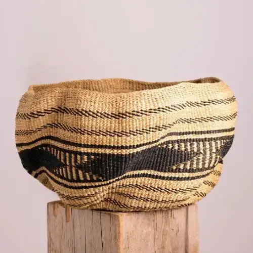 A woven basket with black wave-like patterns sits on a wooden block against a plain light background. The basket has an uneven, organic shape and natural straw color.