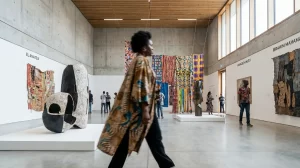 A person in a patterned robe walks through a modern gallery with high ceilings, concrete walls showcasing contemporary African art. Colorful textiles and sculptures displayed throughout the space. Several people observe the artworks.