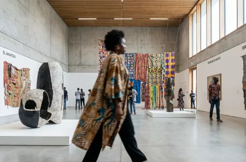 A person in a patterned robe walks through a modern gallery with high ceilings, concrete walls showcasing contemporary African art. Colorful textiles and sculptures displayed throughout the space. Several people observe the artworks.