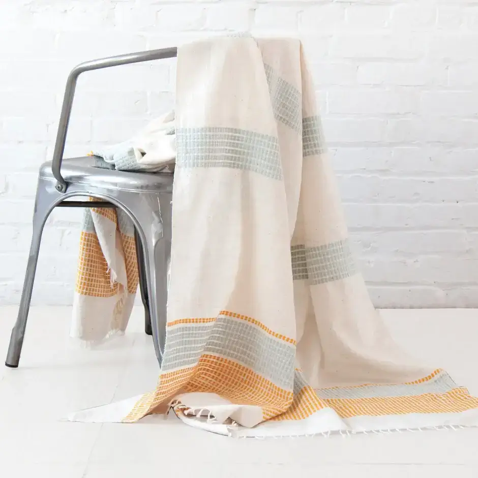A cream-colored woven throw with gray and yellow stripes draped over a metal chair, set against a white brick wall and white floor.