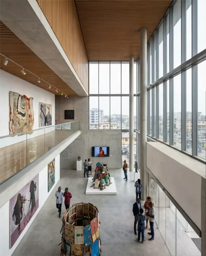A spacious, modern art gallery with high ceilings, large windows, concrete walls, colorful artwork on display, and several people walking and viewing the exhibits. City buildings are visible through the windows.