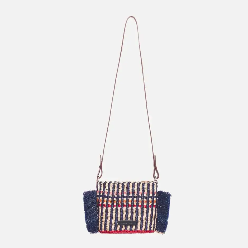 A small woven shoulder bag with a long brown strap, featuring a striped pattern in beige, red, and blue, and blue fringes on the sides.