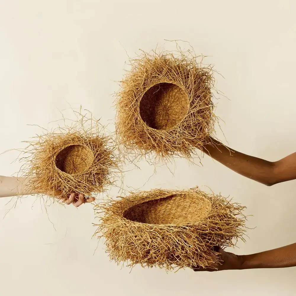 Three hands hold woven baskets with straw-like edges against a plain beige background; each basket varies in size, appearing organic and nest-like.
