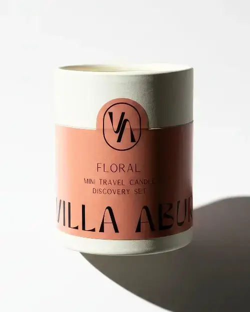 A cylindrical candle container with a pink label that reads FLORAL MINI TRAVEL CANDLE DISCOVERY SET and partially shows the brand name VILLA ABU in black text, placed on a white surface with a shadow.