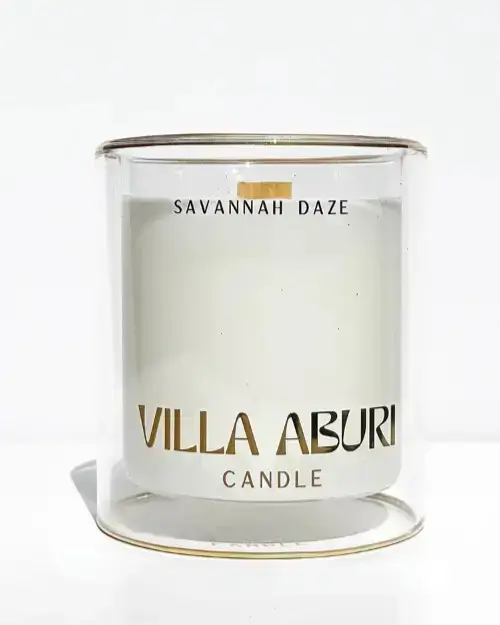 A clear glass candle with white wax inside. The label reads SAVANNAH DAZE at the top and VILLA ABURI CANDLE in gold lettering on the front. The background is plain white.