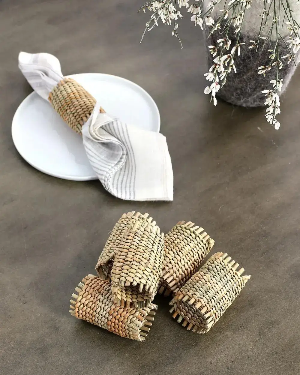 A white plate with a cloth napkin held by a woven napkin ring sits on a table. Four additional woven napkin rings are nearby, and a vase with white flowers is partially visible in the corner.